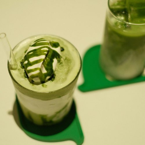 Two iced matcha lattes topped with cream and matcha drizzle on a table.